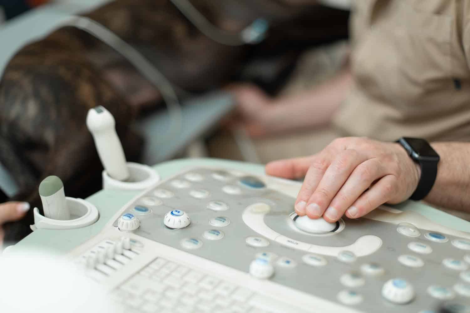 A person operates a veterinary ultrasound machine with a dog lying on the examination table in the background.
